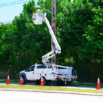 Electrician working on high power lines from a lift on the back of a truck that is parked in the street and surrounded by warning cones